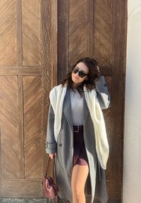 Manteau en laine gris avec col châle blanc, jupe bordeaux à motifs, et un sac à main bordeaux en forme de cœur, le tout sur un fond en bois.