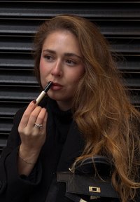 Læbestift i et gyldent og sort etui holdt i en hånd. Modellen har en sort rullekrave og langt, bølget brunt hår. Nærbillede.