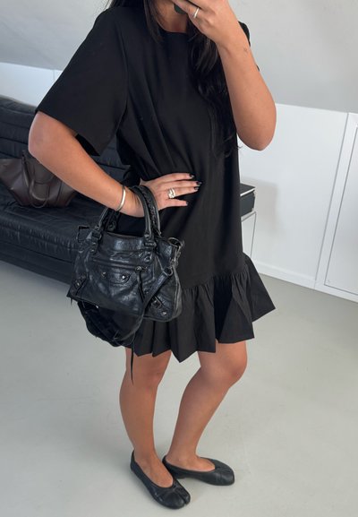 Mujer con vestido negro con volantes que sostiene un bolso de cuero negro, lleva zapatos planos negros con punta partida, de pie en el interior cerca de un sofá negro.