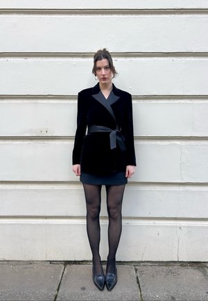 Mujer con el cabello oscuro recogido, que lleva un blazer de terciopelo negro con cinturón de satén, falda negra, medias y zapatos negros puntiagudos, de pie contra una pared blanca.