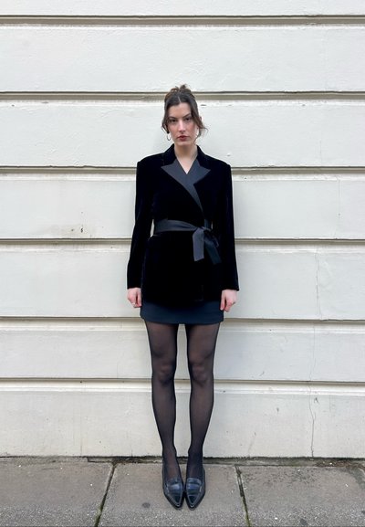 Mujer con el cabello oscuro recogido, que lleva un blazer de terciopelo negro con cinturón de satén, falda negra, medias y zapatos negros puntiagudos, de pie contra una pared blanca.