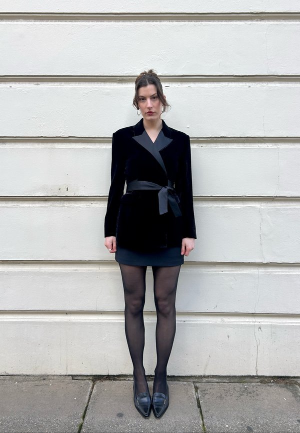 Femme aux cheveux foncés attachés, portant un blazer en velours noir avec ceinture en satin, jupe noire, collants et chaussures noires à bout pointu, debout contre un mur blanc.