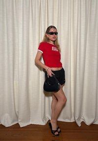 Kvinna i röd Puma-crop-top, svarta shorts, svarta högklackade sandaler och solglasögon, som håller en liten svart handväska, stående framför krämfärgade gardiner.
