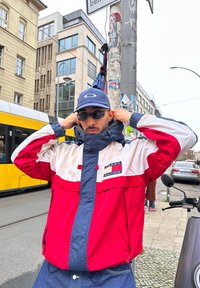 Homme portant une veste Tommy Hilfiger rouge, blanche et bleue, une casquette bleue et des lunettes de soleil, ajustant le col de sa veste dans une rue de la ville.