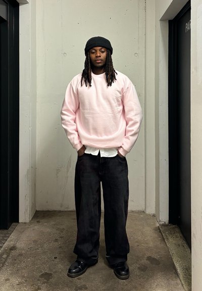 Sudadera rosa claro, camisa blanca debajo, pantalones anchos negros y zapatos negros. Cabello largo y oscuro, de pie en un espacio minimalista.