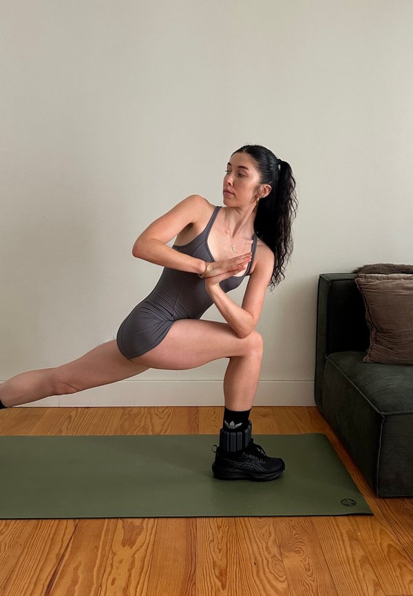 Woman with long dark hair in a grey workout outfit performs a twisted lunge stretch on a green mat in a room with a couch and wooden floor.