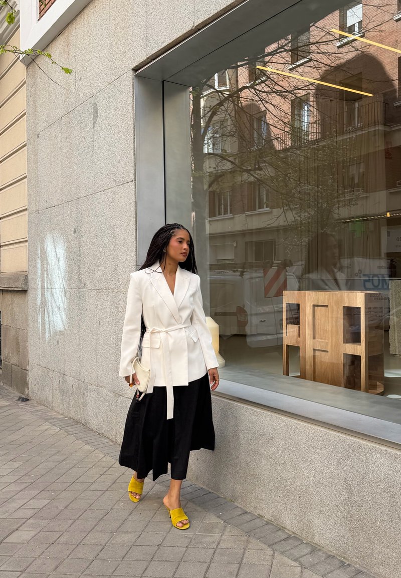 Femme en blazer blanc, jupe noire et sandales jaunes marchant sur le trottoir devant une grande fenêtre en verre montrant du mobilier en bois à l’intérieur.