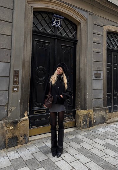 Mujer de cabello largo y rubio, que lleva un abrigo negro, boina y botas, sonríe frente a unas grandes puertas dobles negras con el número 23 Bernardg.