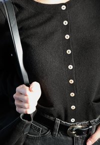 Osoba nosící černý svetr na knoflíky držící popruh černé kabelky, oblečená v tmavých džínách s lesklou černou přezkou opasku.