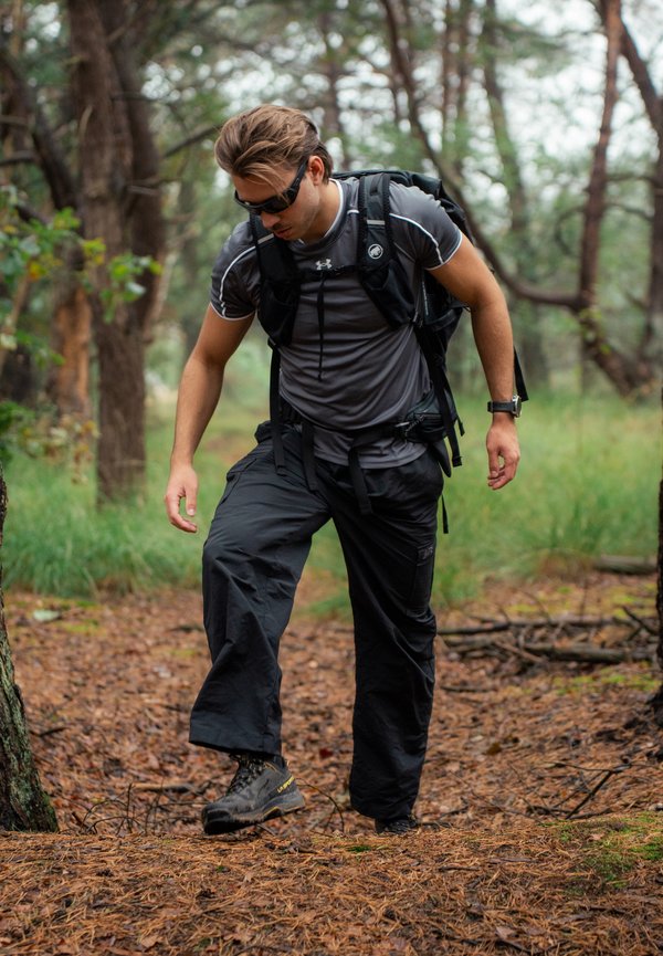 Un hombre con gafas de sol y una mochila camina por un sendero cubierto de agujas de pino que atraviesa un bosque.