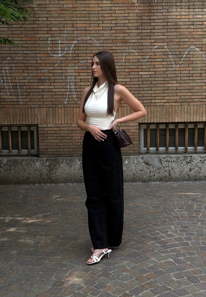 Jeune femme aux longs cheveux foncés se tient dans une rue pavée, portant un haut blanc sans manches, un pantalon noir et des sandales blanches à talons.
