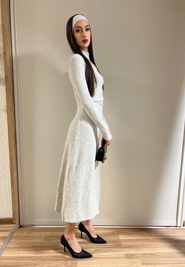 Femme portant une robe gris clair à manches longues, des talons hauts noirs, un bandeau blanc, tenant un petit sac à main noir, debout à l'intérieur contre un mur uni.