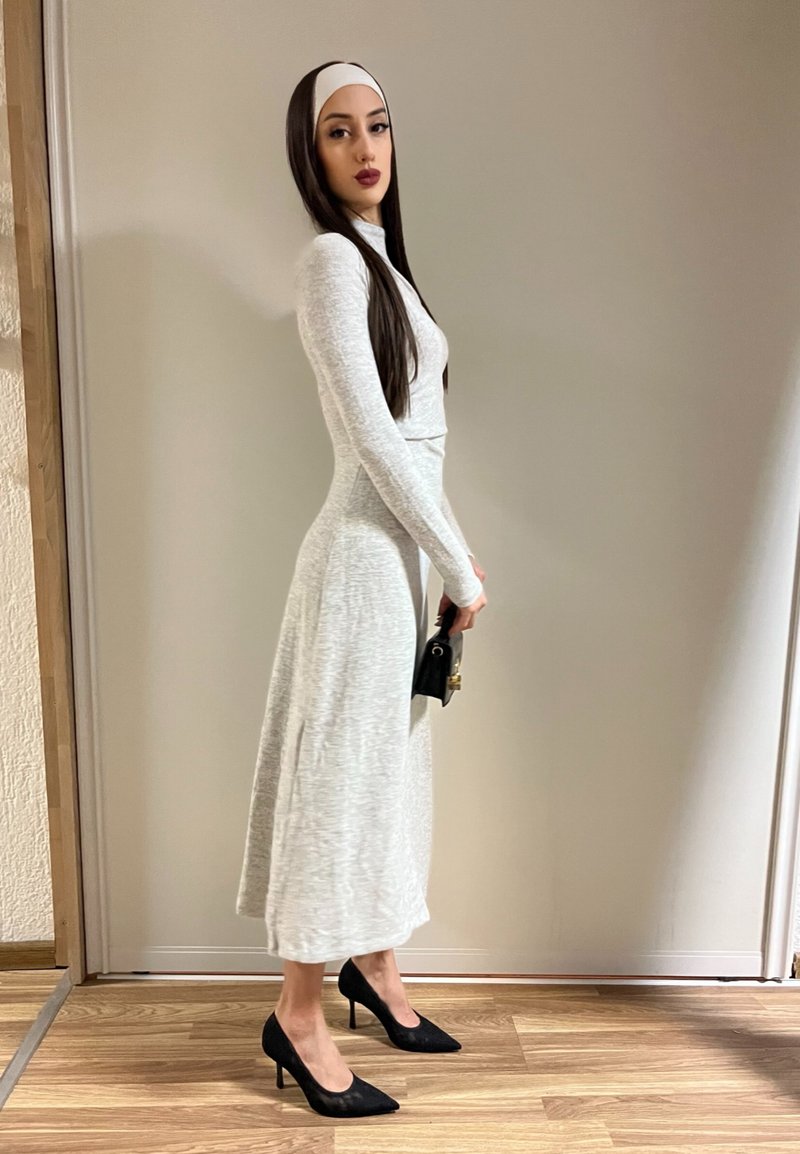 Femme portant une robe gris clair à manches longues, des talons hauts noirs, un bandeau blanc, tenant un petit sac à main noir, debout à l'intérieur contre un mur uni.