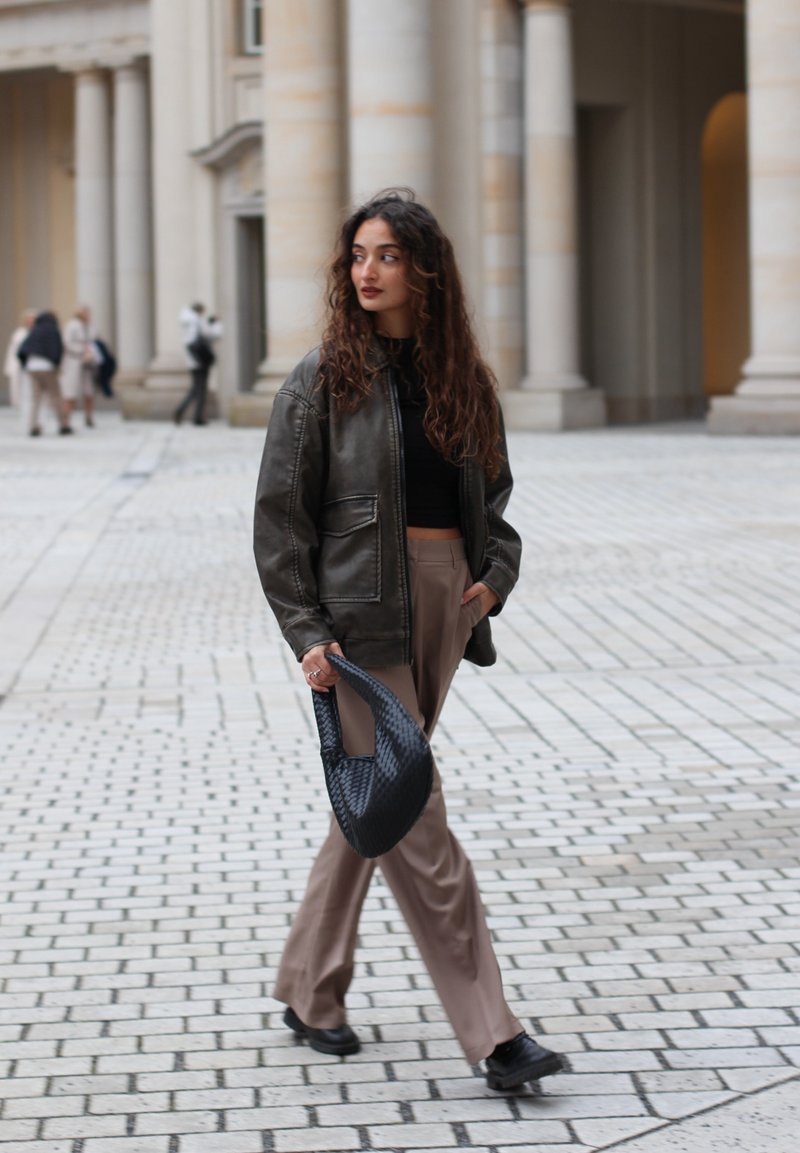 Σκοτεινός καφέ δερμάτινος μπουφάν με ορατές ραφές, μαύρο κοντό τοπ, ανοιχτό μπεζ παντελόνι με φαρδιές ζωές και μια υφασμάτινη μαύρη τσάντα. Φόντο από κυβόλιθους.