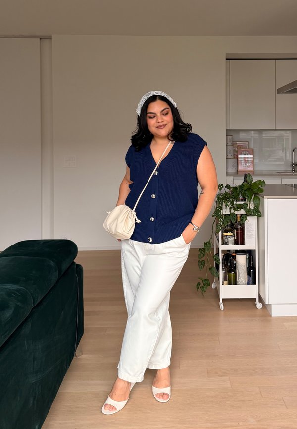 Vrouw met donker haar die een marineblauw mouwloos topje met knopen draagt, witte broek, witte sandalen, hoofddoek en schoudertas, staand in een moderne keuken.