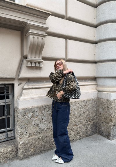 Chaqueta de estampado de leopardo, jeans de pierna ancha de color azul oscuro y mules blancos con tachuelas. Sosteniendo un bolso negro con tachuelas plateadas. Fondo urbano.