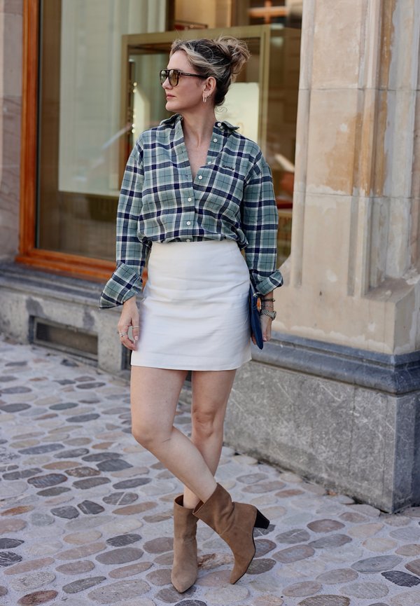 Frau steht auf gepflasterter Straße, trägt grünes kariertes Hemd, weißen Minirock, braune Stiefeletten, Sonnenbrille und hält eine Clutch-Tasche.