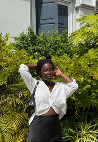 Kvinne med briller og hvit skjorte bundet i midjen står med hendene bak hodet foran grønne, bladete planter og en blå dør.