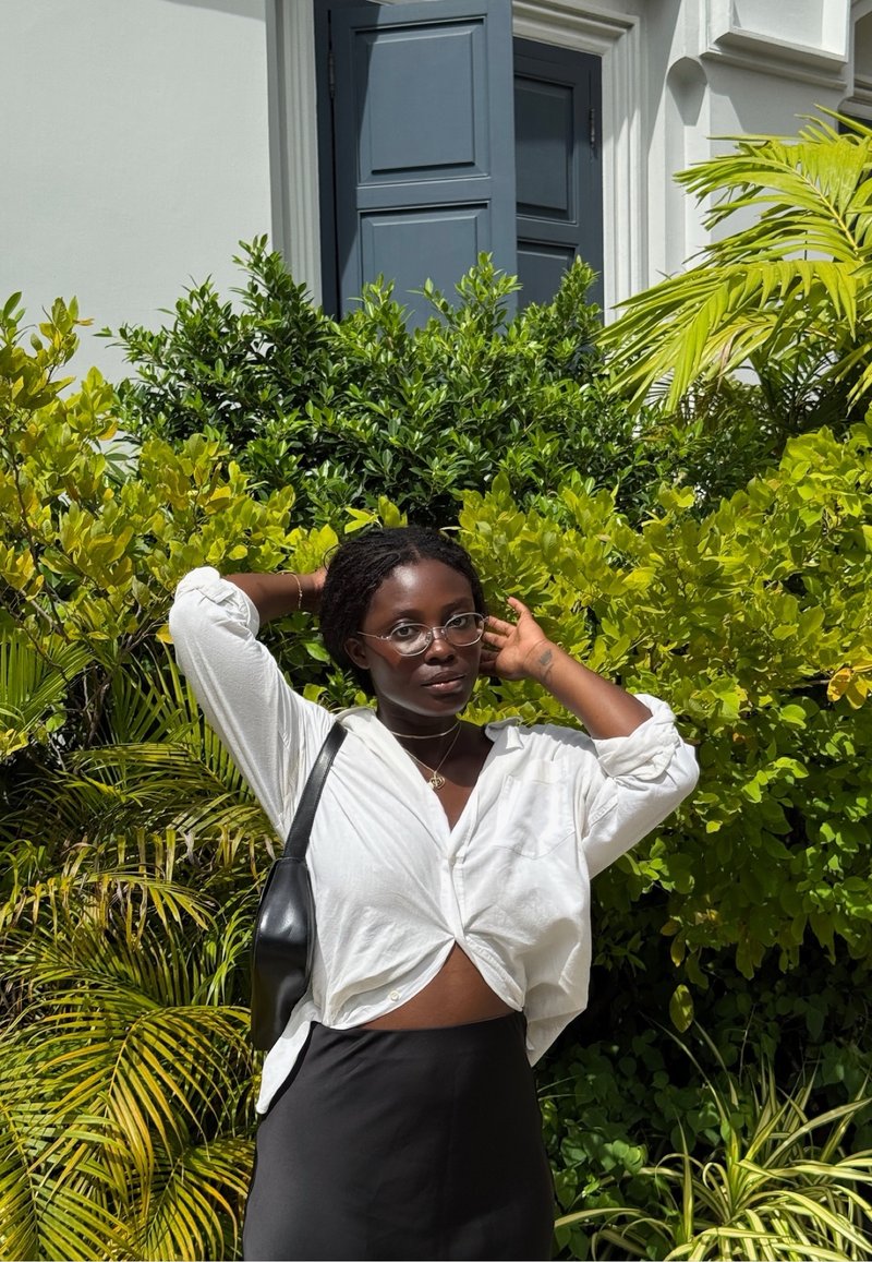 Vrouw met een bril en een wit shirt dat om de taille is geknoopt, staat met haar handen achter haar hoofd voor groene bladeren en een blauwe deur.