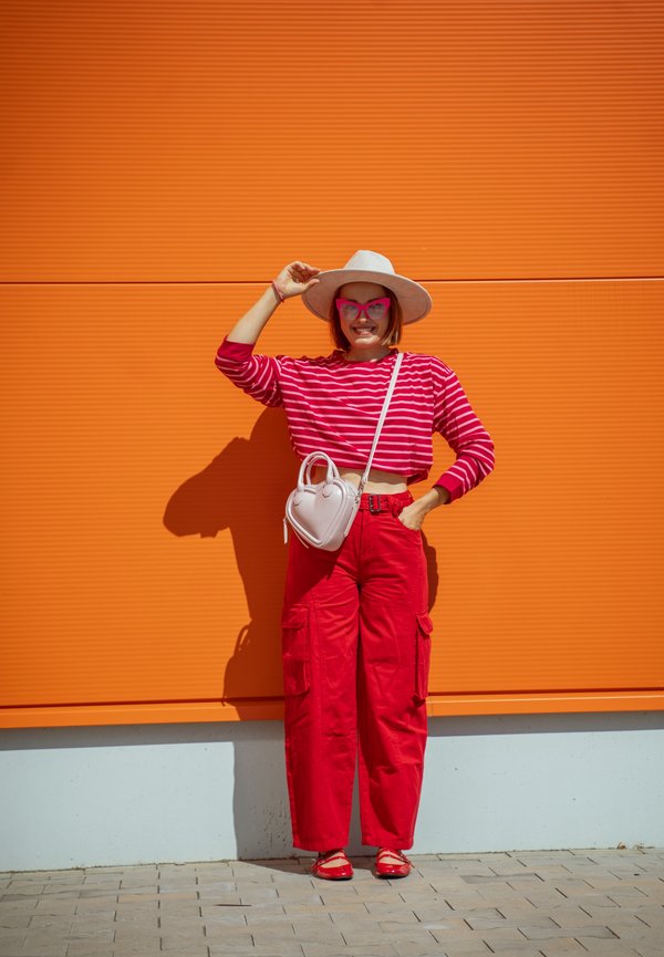 Mujer con camisa de rayas rojas, pantalones rojos, sombrero blanco y gafas rosas, de pie contra una pared naranja sosteniendo el ala del sombrero con una mano.