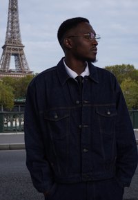 Jeansjacka med knäppning, mörkblå färg, två fickor fram, bärs över en vit skjorta och svart slips. Eiffeltornet i bakgrunden.