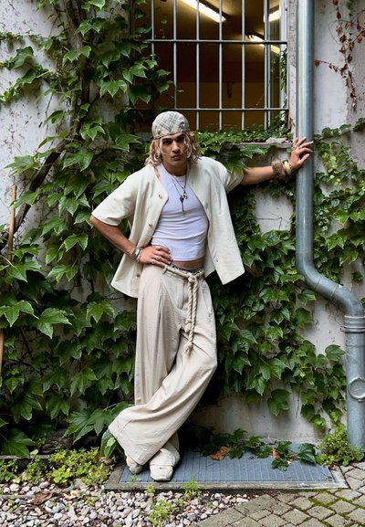 Camisa corta de manga corta crema sobre una camiseta blanca, combinada con pantalones sueltos de color beige claro con un cinturón de cuerda, frente a una pared cubierta de enredaderas.