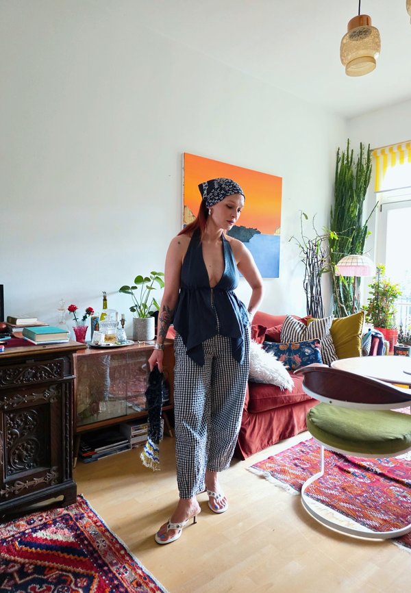 Donna che indossa un top nero e pantaloni a fantasia si trova in un soggiorno colorato con piante, mobili vintage e un'opera d'arte luminosa sulla parete.