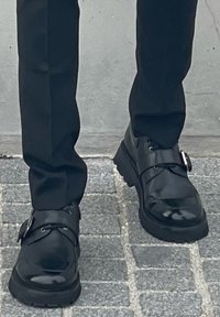 Μαύρες δερμάτινες παπούτσια με γυαλιστερή εμφάνιση, που διαθέτουν μια φαρδιά ζώνη και στρογγυλή αγκράφα, ανθεκτική σόλα και λεπτομέρειες με κορδόνια.