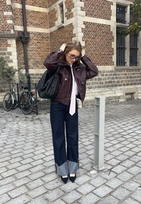 Vinröd skinnjacka, ljusrosa slips, mörka jeans med fransig fåll, svart vävd tygväska, svarta högklackade skor, gatstensbakgrund.