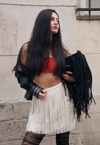 Top corto rosso a costine abbinato a una gonna bianca con frange. L'outfit è completato da una giacca di pelle nera con borchie argentate e leggings con fantasia.