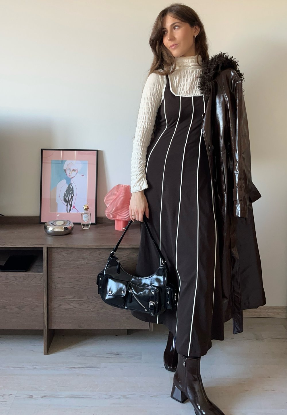 Abito nero con righe verticali bianche, dolcevita bianco strutturato, cappotto lungo marrone lucido e borsa nera in vernice.