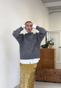 Ohlapen sivi pleteni pulover z visokim ovratnikom, nošen čez belo srajco. Bleščeča zlata krilo s sequinami sega do tal. Preprosto notranje ozadje.