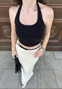 Black halter neck crop top paired with a long, light beige skirt. Silver cuff bracelet, black handbag, and flat shoes visible. Smooth textures and a simple design.