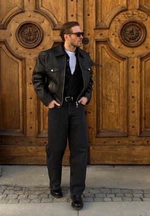 Hombre con chaqueta de cuero negra, gafas de sol y vaqueros negros, de pie con las manos en los bolsillos frente a una gran puerta de madera ornamentada.