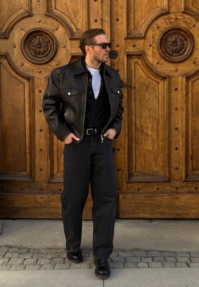Hombre con chaqueta de cuero negra, gafas de sol y vaqueros negros, de pie con las manos en los bolsillos frente a una gran puerta de madera ornamentada.