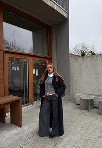 Jeune femme portant des lunettes, un long manteau noir, un cardigan gris raccourci et un pantalon large, debout devant un bâtiment moderne avec une porte en verre et des murs en béton.