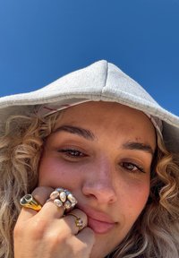 Grå hettegenser med en myk tekstur, omgitt av krøllete hår. Flere ringer på fingrene: gull, perlelignende, og et lite stjernedesign. Blå himmel i bakgrunnen.