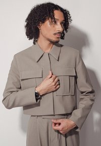Veste coupée gris clair avec de grandes poches avant, un large col et des manches longues. Tissu lisse avec des détails de couture précis.