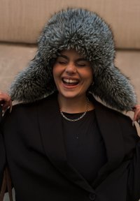 Falsk pelsfellelue i grå toner, med lange klaffer og tykk, myk tekstur. Brukt med en svart blazer og lagdelt kjedekjede.