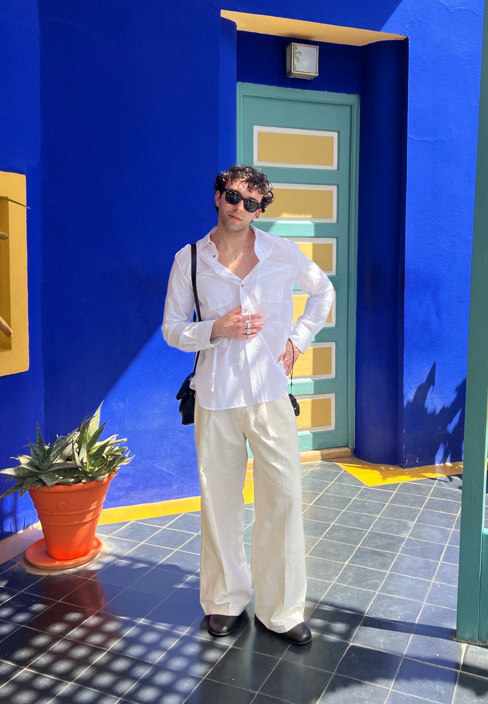 Camicia bianca a maniche lunghe, pantaloni crema a gamba larga, occhiali da sole neri e borsa nera. In piedi su piastrelle decorate vicino a una pianta in vaso.