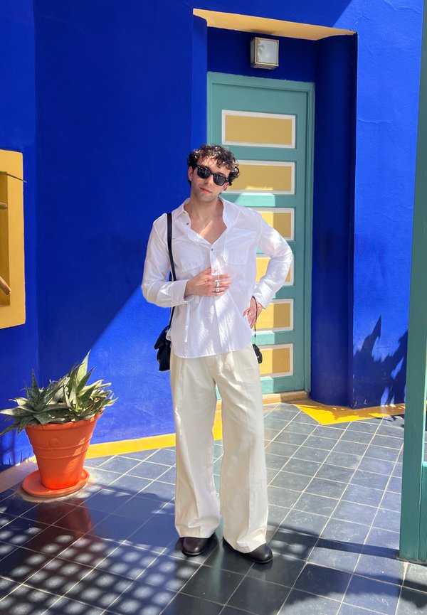 Camicia bianca a maniche lunghe, pantaloni crema a gamba larga, occhiali da sole neri e borsa nera. In piedi su piastrelle decorate vicino a una pianta in vaso.