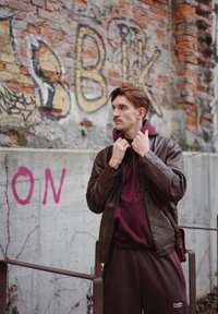 Chaqueta de cuero marrón con acabado texturizado, superpuesta sobre una sudadera con capucha burdeos y pantalones deportivos a juego. El fondo presenta ladrillos envejecidos y grafiti.