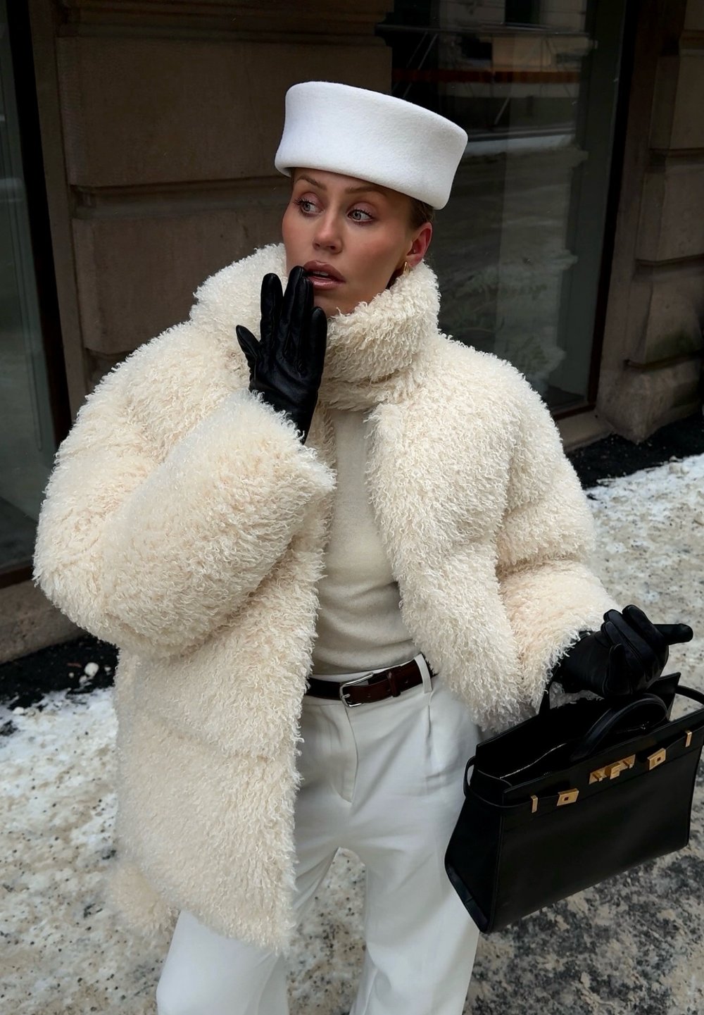 Femme en manteau en fourrure crème, chapeau blanc, gants en cuir noir tenant un sac à main noir sur un trottoir urbain enneigé.