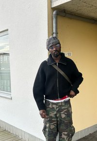 Suéter preto canelado com zíper, calças de camuflagem, gorro cinza e óculos. De pé contra uma parede amarelo-claro com um tubo.