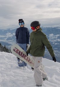 To personer i snøfylt terreng, en i grønn jakke og svarte hansker som holder et hvitt snowboard med rødt tekst, den andre i blå pufferjakke.