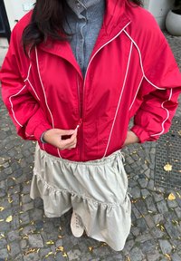 Rød vindjakke med hvide striber og lynlåslukning foran, kombineret med et lyst beige lagdelt skørt og en grå ribbet top. Afslappede sneakers er synlige.