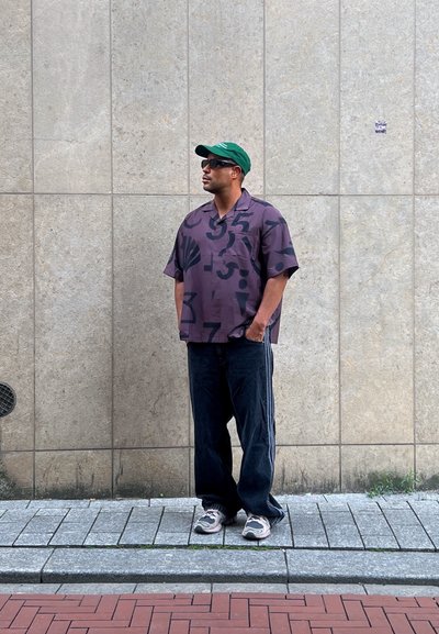 Camisa de manga corta morada con un patrón gráfico, jeans de pierna ancha azul oscuro y zapatillas grises. Una gorra verde y unas gafas de sol completan el conjunto.