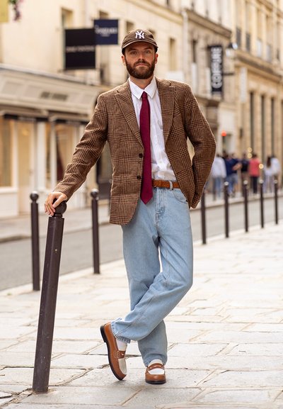 Hombre vestido con un blazer de cuadros marrón, camisa blanca, corbata roja, jeans azul claro, zapatos marrones y una gorra oscura, apoyándose en un poste de la calle en la acera de la ciudad.