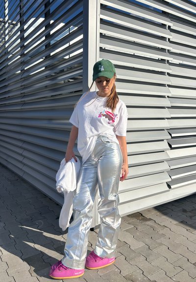 Pantalones plateados, brillantes y de cintura alta combinados con una camiseta gráfica blanca. Una gorra verde y unos zapatos de plataforma rosas completan el conjunto. Estilo urbano casual.