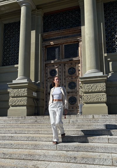 Blazer de lino blanco y pantalones a juego combinados con un top corto blanco. Puerta de madera oscura con intrincadas tallas y escalones de piedra.